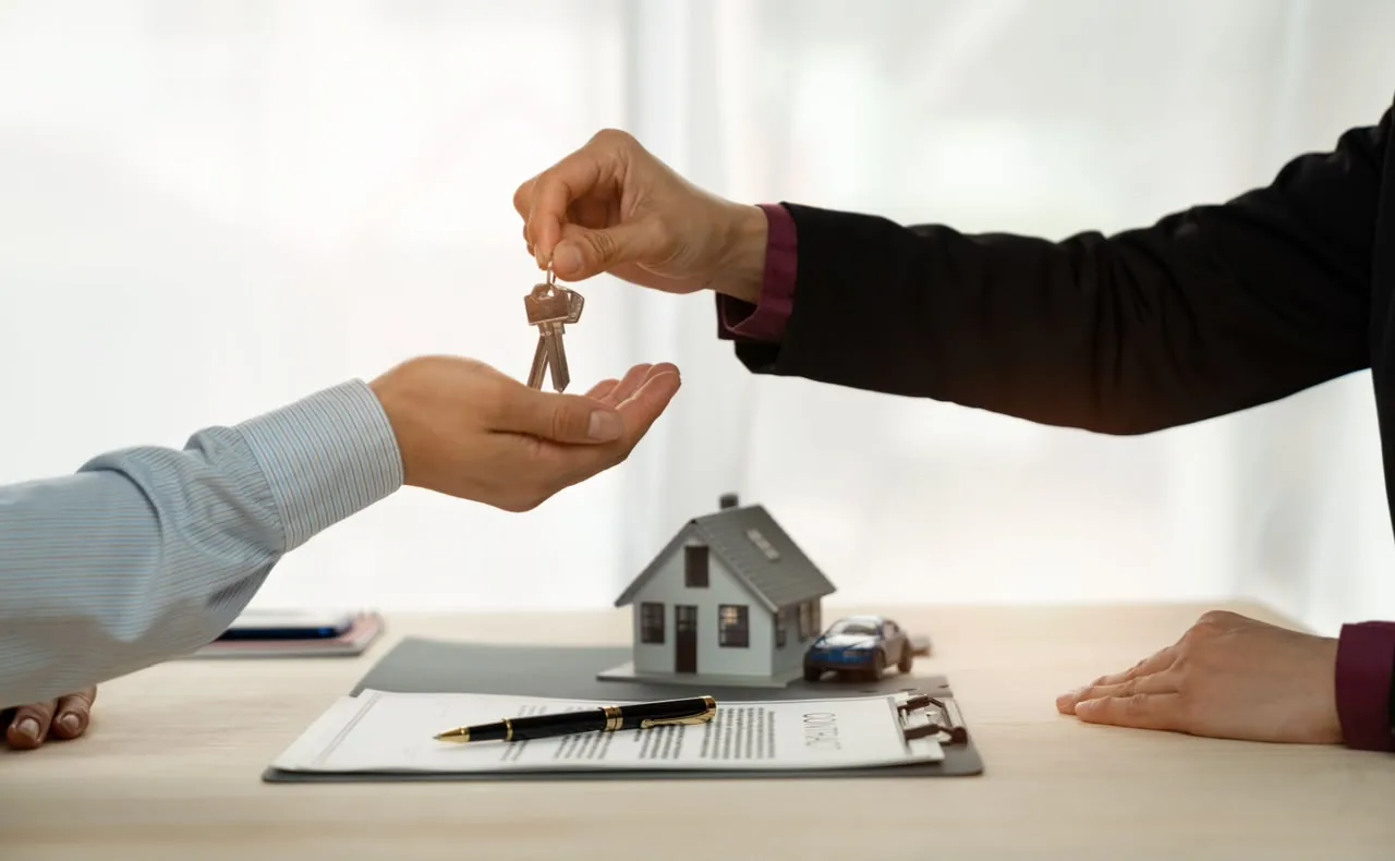 House keys being handed over across a desk with a model house and a contract